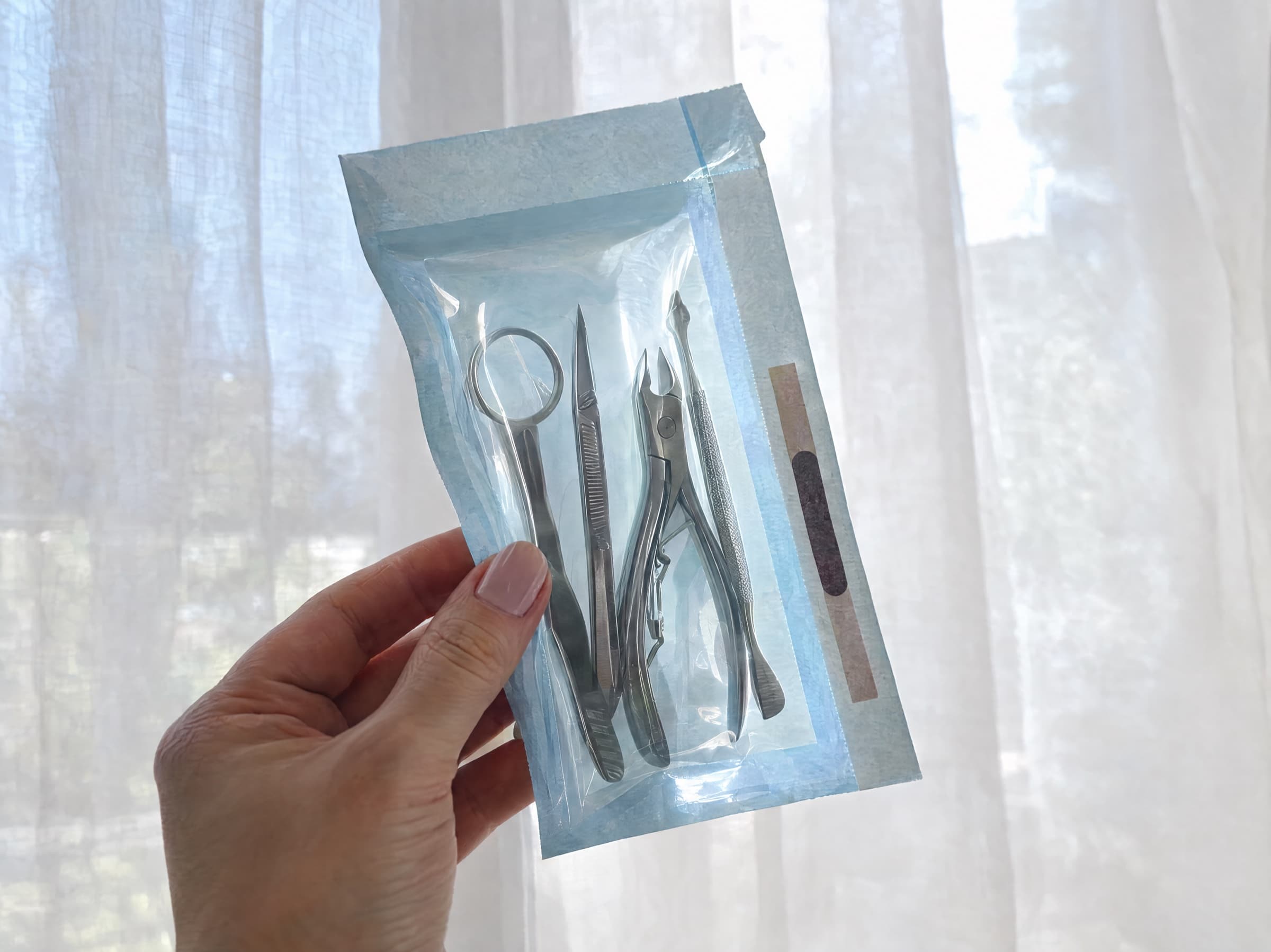 A sealed sterilisation pouch of metal manicure tools held up to the light so its indicator strip is visible